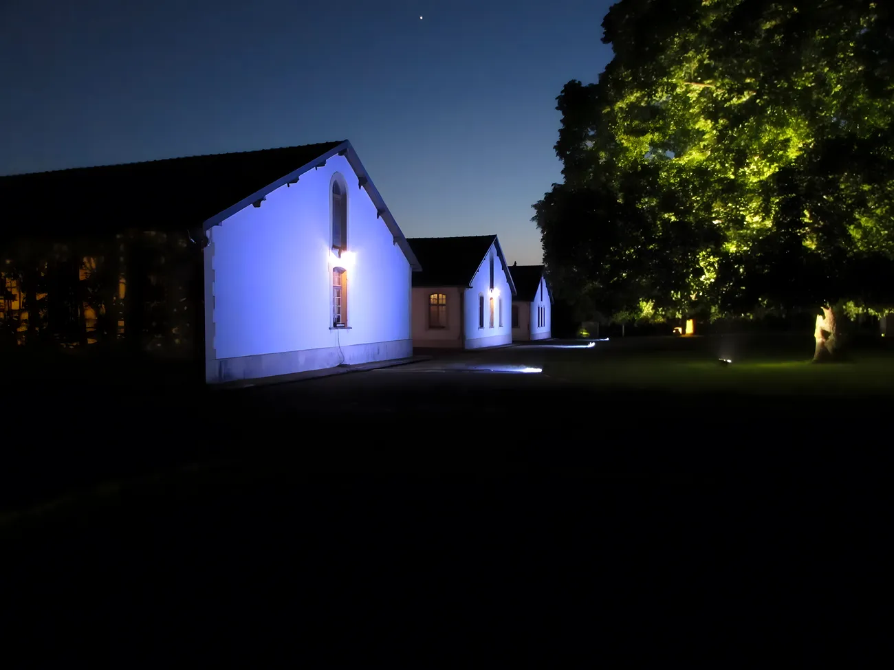 The memorial barracks at night, lit in deep blue with a single gold-lit tree